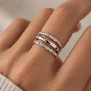 以下が含まれることがあります： 3つの異なる列で構成されたシルバー調のマルチバンドリング。上下のバンドには小さな透明の輝く石が埋め込まれており、中央のバンドは滑らかで光沢のあるシルバー調の金属面になっています。