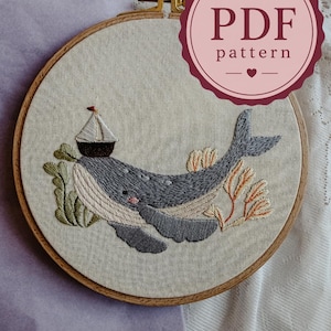 Puede incluir: Un bastidor de bordado con una pieza de tela que muestra una ballena gris bordada, un pequeño velero sobre su lomo, algas verdes y coral naranja. El texto PDF pattern aparece en una insignia circular de color rojo oscuro arriba a la derecha.