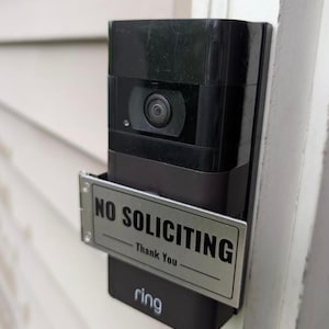 May include: A black video doorbell device mounted on a wall, featuring a rectangular silver metal sign attached to its front. The sign displays the black text NO SOLICITING followed by the text Thank You with horizontal lines above and below.