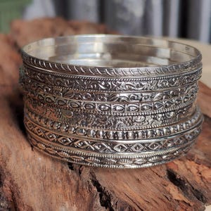Könnte beinhalten: Ein Stapel aus sieben silberfarbenen Metallarmreifen, von denen jeder ein eigenes geprägtes Muster aufweist. Die Designs umfassen komplexe florale Motive, geometrische Formen, perlenartige Texturen und lineare Gravuren für ein abwechslungsreiches Aussehen.