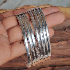 Peut inclure: Un ensemble de cinq bracelets joncs argentés polis avec une surface lisse et arrondie. Les bracelets sont disposés en pile, présentant une finition métallique réfléchissante qui capte la lumière. Le texte LiyanshCollection27 est visible.