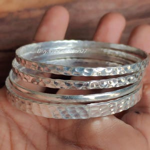 Peut inclure: Un ensemble de trois bracelets joncs argentés avec une finition martelée et texturée. Les bracelets sont empilés ensemble, présentant une surface métallique réfléchissante. Le texte DivyanshCollection27 est visible sur les bracelets.