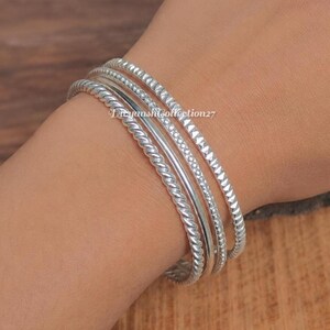 Può includere: Un set di quattro bracciali rigidi color argento con texture distinte. La collezione include un bracciale a corda intrecciata, uno liscio e lucido, uno con perline e uno con un motivo geometrico a forma di piramide.