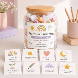 May include: A clear glass jar with a bamboo lid filled with colorful candies and white cards. The label reads Mutmacherkarten ZUM SCHULSTART 50 Karten für kleine Herzen mit großen Träumen. Eight individual cards with illustrations and German text are displayed in front.