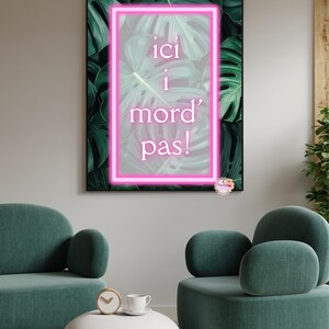Könnte beinhalten: Ein rechteckiges Wandbild mit einem leuchtend pinken Neon-Rahmen um den französischen Text 'ici i mord pas!', platziert vor einem Hintergrund aus dunkelgrünen Monstera-Blättern. Das Bild ist in einem dünnen schwarzen Rahmen gefasst.