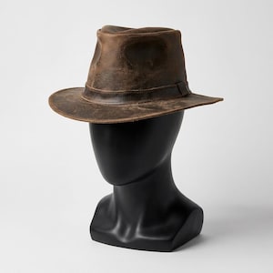 Puede incluir: Un sombrero fedora de cuero marrón oscuro con un acabado desgastado y envejecido. El sombrero presenta una copa pellizcada y un ala estructurada, acentuado por una banda de cuero a juego alrededor de la base de la copa con costuras visibles.
