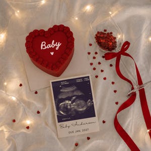 Könnte beinhalten: Ein herzförmiger roter Kuchen mit weißer Schrift Baby und einem kleinen Herz, ein Glas gefüllt mit roten herzförmigen Konfetti und einer roten Schleife sowie ein rechteckiger Ultraschallausdruck mit der Aufschrift Baby Anderson DUE JAN. 2027.