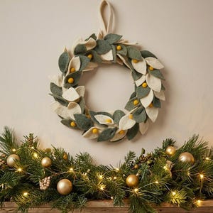 Peut inclure: Une couronne circulaire composée de feuilles en feutrine superposées dans des tons vert sauge et crème, agrémentée de petites boules en feutrine jaune moutarde. La couronne présente une boucle en tissu sur le dessus pour la suspendre.