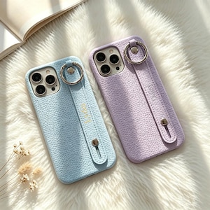 May include: Two textured smartphone cases, one light blue and one lavender, each featuring a vertical strap with a circular metal ring attachment. The cases have cutouts for a triple-lens camera system and a metallic stud securing the strap.