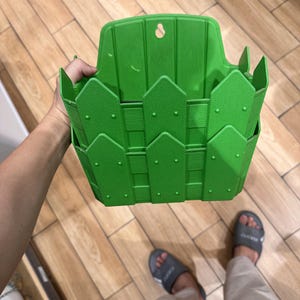 Könnte beinhalten: Ein leuchtend grüner Wandpflanzkübel aus Kunststoff mit einem dekorativen Lattenzaun-Design. Der Behälter verfügt über eine flache Rückwand mit einer Schlüssellochöffnung zum Aufhängen und eine texturierte Oberfläche in Holzoptik.