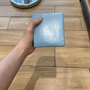 Könnte beinhalten: Eine quadratische, hellblaue Keramikfliese mit glatter, glänzender Oberfläche. Die Fliese hat leicht abgerundete Kanten und eine gleichmäßige, einfarbige Oberfläche, die das Licht reflektiert und ein klares, schlichtes Design aufweist.