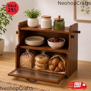 May include: A two-tier wooden bread box with a drop-down fluted glass door. The top shelf holds a white potted plant, a white ceramic jar, and a green glass jar. The bottom shelf contains a loaf of bread, bagels, and croissants in paper bags.