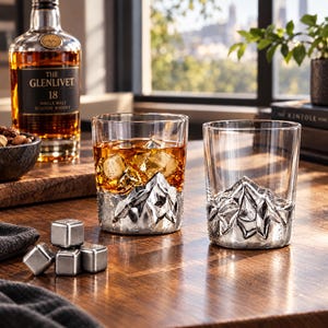 May include: Two clear glass tumblers featuring a sculpted, metallic mountain range design at the base. One glass is filled with amber liquid and ice cubes, while the other is empty. Four metallic cooling cubes sit nearby on the wooden surface.