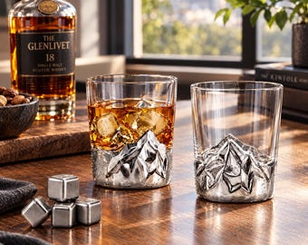 May include: Two clear glass tumblers featuring a sculpted, metallic mountain range design at the base. One glass is filled with amber liquid and ice cubes, while the other is empty. Four metallic cooling cubes sit nearby on the wooden surface.