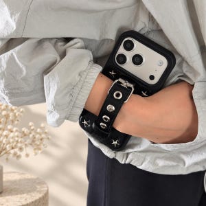 Puede incluir: Una funda de smartphone negra con una correa de muñeca integrada, hebilla plateada y ojales metálicos. La funda está decorada con pequeños remaches plateados en forma de estrella y tiene un recorte para las lentes de la cámara trasera.