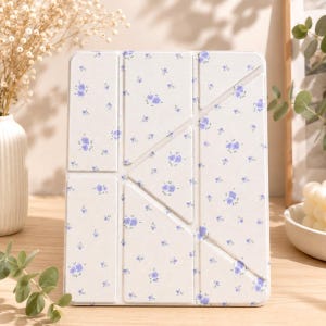 May include: A rectangular tablet case featuring a light cream background with a repeating pattern of small purple floral clusters. The case has a segmented, foldable design with visible creases that allow for multiple standing positions.