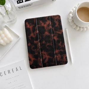 Op de afbeelding: Een rechthoekige tablethoes met een donkerbruin en zwart schildpadpatroon en een drievoudig vouwontwerp. De hoes heeft een gladde, matte afwerking en is verticaal op een plat oppervlak geplaatst naast een witte styluspen.