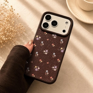 May include: A dark brown smartphone case featuring a textured fabric-like surface with a repeating pattern of small, colorful embroidered flowers. The case includes a raised, integrated camera lens protector in a matching dark brown frame.