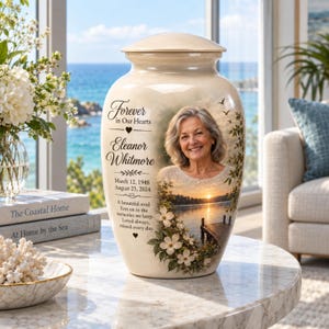 Op de afbeelding: Een crèmekleurige keramische urn met het portret van een glimlachende vrouw, een zonsondergang bij een meer met een houten steiger en witte kornoeljebloemen. De tekst luidt: Forever in Our Hearts, Eleanor Whitmore, 12 maart 1948, 25 augustus 2024.