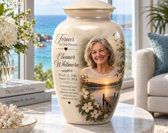 Puede incluir: Una urna funeraria de cerámica color crema con el retrato de una mujer sonriente, una escena de lago al atardecer con un muelle de madera y flores de cornejo blancas. El texto dice: Forever in Our Hearts, Eleanor Whitmore, 12 de marzo de 1948, 25 de agosto de 2024.