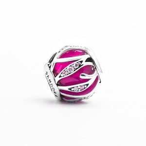 May include: A spherical silver charm bead featuring a vibrant hot pink enamel inlay with a cut-out leaf-like pattern. The silver overlay is accented with small, clear, round-cut stones. The word PANDORA is engraved on the side of the silver frame.