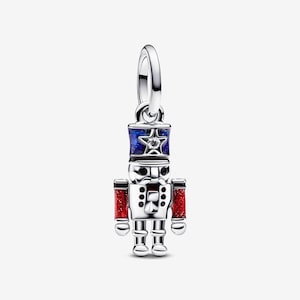 May include: A silver nutcracker charm featuring a blue enamel hat with a central star and a clear stone, black enamel eyes and mustache details, and red glitter enamel accents on the arms. The charm includes a circular bail for attachment.