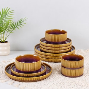 May include: A set of ceramic dinnerware featuring a ribbed exterior in a mustard yellow glaze and a smooth, deep purple interior. The collection includes stacked plates and bowls with a consistent vertical texture and a glossy finish throughout.