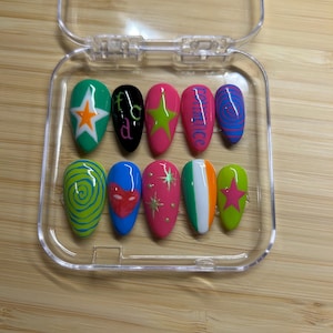 Pode incluir: Um conjunto de dez unhas postiças em formato amêndoa num estojo de plástico transparente. Os designs incluem uma unha verde com estrela laranja, uma preta com texto verde, uma rosa com estrela verde, uma rosa com a palavra romance e uma azul com espirais roxas. Outras unhas apresentam espirais verdes sobre amarelo, um coração vermelho sobre azul, estrelas douradas sobre rosa, uma faixa tricolor vertical e uma estrela rosa sobre verde lima.