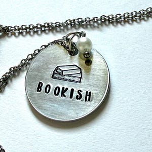 May include: A silver-toned circular pendant necklace featuring a stamped graphic of two stacked books above the word BOOKISH in black lettering. The pendant is attached to a silver chain and includes a small, round, white pearl-like bead accent.