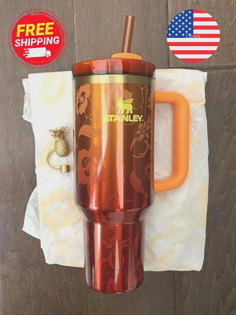 May include: A metallic orange Stanley tumbler featuring a floral pattern, a matching orange handle, a lid with an orange straw, and a small gold-toned pineapple charm accessory attached to the side of the cup near the top rim.