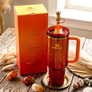 May include: A red Stanley Quencher H2.0 FlowState tumbler with a gold pineapple-shaped straw topper, featuring a matching orange box labeled Stanley Karol G. The tumbler has a handle, a clear lid, and a metallic finish with floral patterns.