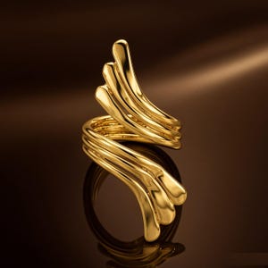 Op de afbeelding: Een goudkleurige ring met een bypass-ontwerp met twee overlappende, waaiervormige secties. Elke sectie bestaat uit drie gladde, afgeronde, parallelle banden die naar buiten buigen, wat zorgt voor een vloeiend, gelaagd uiterlijk met een glanzende afwerking.