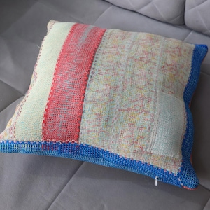 Peut inclure: Un coussin carré au design patchwork composé d'une bande verticale rouge, d'une section beige clair aux motifs multicolores délavés et d'une bordure bleue unie sur les côtés. Le tissu présente un tissage texturé visible.