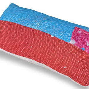 Peut inclure: Un coussin rectangulaire présentant un motif en patchwork avec des sections vives de tissu bleu clair, rouge et violet. La surface affiche un motif dense de fines coutures blanches, avec un empiècement rectangulaire rose sur le côté droit.
