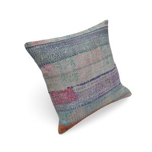 May include: A square throw pillow featuring a patchwork design of faded fabric strips in shades of light blue, grey, and white, accented with small rectangular patches of pink, purple, and orange, finished with visible running stitch details.
