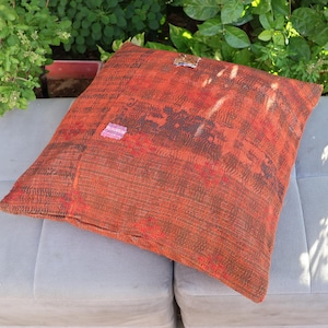 May include: A square throw pillow featuring a textured, multi-tonal orange and dark brown fabric with visible running stitch patterns throughout. The surface includes a small, distinct patch of light pink fabric and a small dark fabric accent.