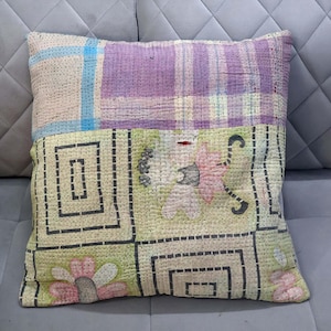 Peut inclure: Un coussin carré au design patchwork avec des surpiqûres apparentes. La partie supérieure présente des motifs à carreaux violets et crème, tandis que la partie inférieure affiche des carrés vert clair avec des bordures géométriques noires et des motifs floraux roses.