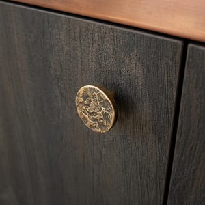 Puede incluir: Un pomo de armario circular con un acabado de latón martillado y texturizado fijado a una puerta de armario de madera oscura. Sobre la puerta hay una moldura horizontal lisa de color cobre que recorre el ancho del marco.