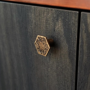 Puede incluir: Un pomo de armario hexagonal con un acabado de latón martillado y texturizado, fijado a una puerta de armario de madera de color gris oscuro con un patrón de veta visible. El pomo presenta un aspecto metálico envejecido con hendiduras.