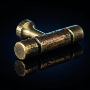 Op de afbeelding: Een T-vormige meubelknop van antiek messing met een getextureerde, gehamerde afwerking. De horizontale balk heeft een cilindrische vorm met een centrale groef, terwijl de verticale steel verbonden is met een ronde basis.