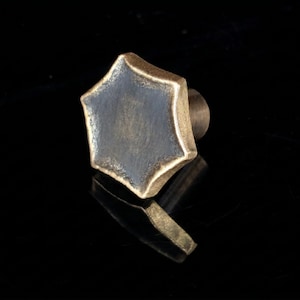 Puede incluir: Un tirador de mueble hexagonal con una superficie central oscura y texturizada, bordeada por un acabado metálico en tono dorado. El tirador cuenta con una base cilíndrica en la parte posterior, diseñada para fijarse a puertas y cajones.