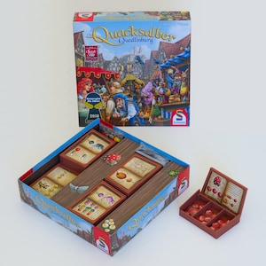 Könnte beinhalten: Ein Brettspiel namens Die Quacksalber von Quedlinburg mit einer Schachtel, die eine mittelalterliche Marktszene zeigt, einer offenen Spielschachtel mit Fächern für Spielkarten und einer kleinen braunen Ablage mit roten Spielsteinen.