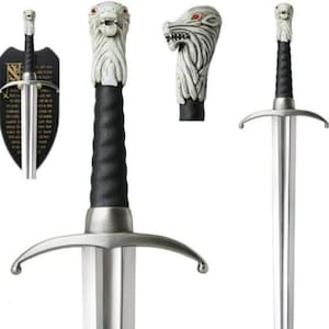May include: A metal sword featuring a pommel shaped like a white wolf head with red eyes, a black textured grip, and a silver crossguard. The sword is displayed alongside a black shield-shaped wall mount with printed text and a separate wolf head.