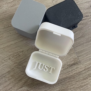 Könnte beinhalten: Drei kleine rechteckige Aufbewahrungsboxen mit Klappdeckeln. Eine Box ist weiß und geöffnet, mit dem Wort JUST auf dem Innenboden eingeprägt. Die anderen beiden Boxen sind geschlossen, eine in Hellgrau und die andere in mattem Schwarz.