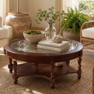 May include: A round wooden coffee table with a glass top and a lower shelf, positioned in a living room with a sofa, two rattan armchairs, a large potted olive tree, a fern, and books stacked on the table surface.