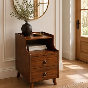 May include: A dark brown wooden nightstand featuring two drawers with circular metal ring pulls, an open storage shelf, and four tapered legs. A black vase with green foliage and a small gold dish sit on the top surface above two books.