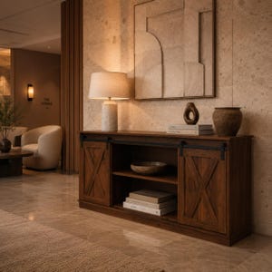 May include: A dark brown wooden console table featuring two sliding barn-style doors with an X-pattern design, a central open shelf, and black metal hardware. The top surface holds a stone-textured table lamp, a dark vase, and stacked books.