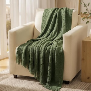 Peut inclure: Un jeté de lit en tricot vert sauge avec un motif en losanges texturé et des bords à franges, drapé sur un fauteuil rembourré de couleur crème. La couverture présente un design tissé uniforme et retombe sur le devant du fauteuil.