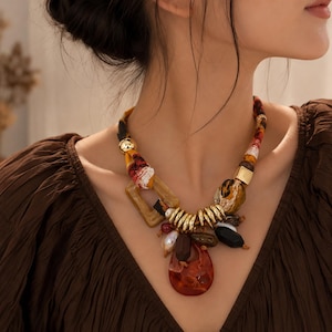 Op de afbeelding: Een statement ketting met een met stof omwikkeld koord in warme tinten, geaccentueerd met goudkleurige metalen kralen, een vierkante hars schakel en een verzameling diverse bedels, waaronder een grote doorschijnende barnsteenkleurige hanger.