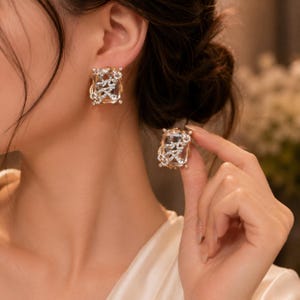 May include: A pair of rectangular stud earrings featuring a large, clear, faceted central stone surrounded by a delicate, gold-toned metal frame adorned with small, sparkling white crystals arranged in an intricate, swirling vine-like pattern.